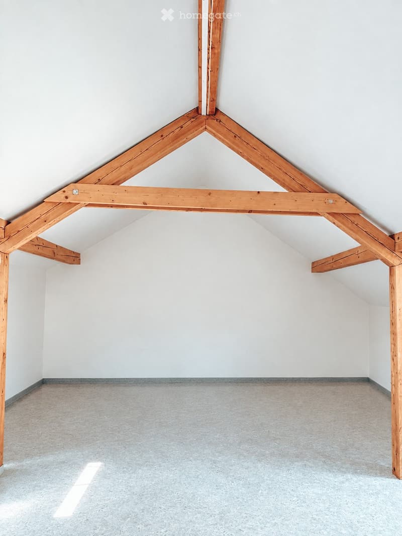 Empty attic space with exposed wooden beams and white walls, featuring a sloped ceiling and grey carpeted floor.
