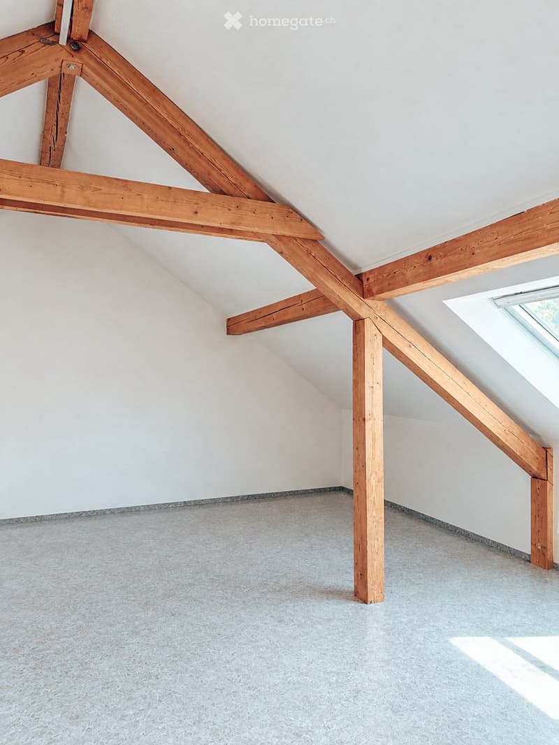 Empty attic space with exposed wooden beams, white walls, and a skylight window allowing natural light.