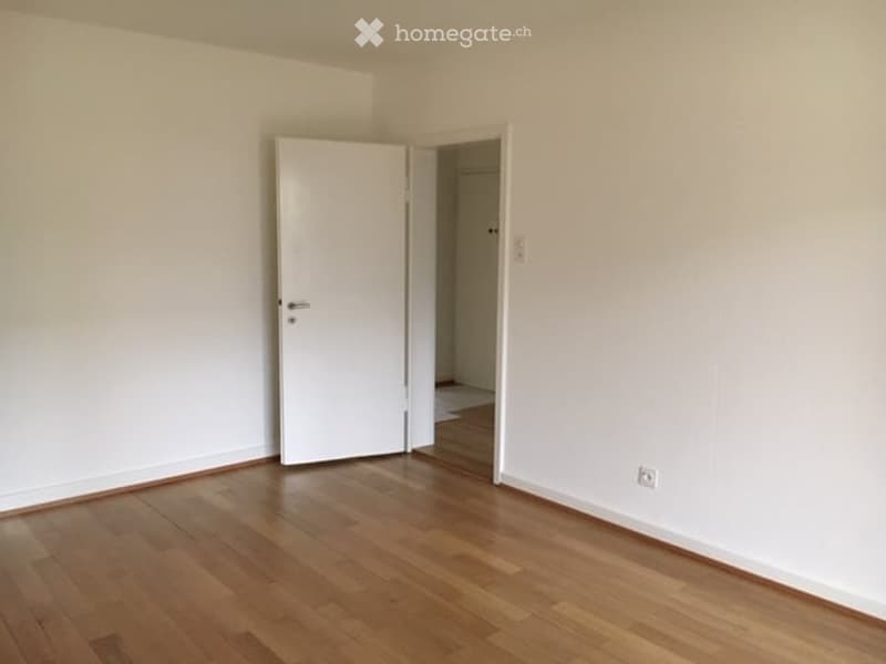 Empty room with wooden flooring and white walls, featuring a partially open white door leading to another room.