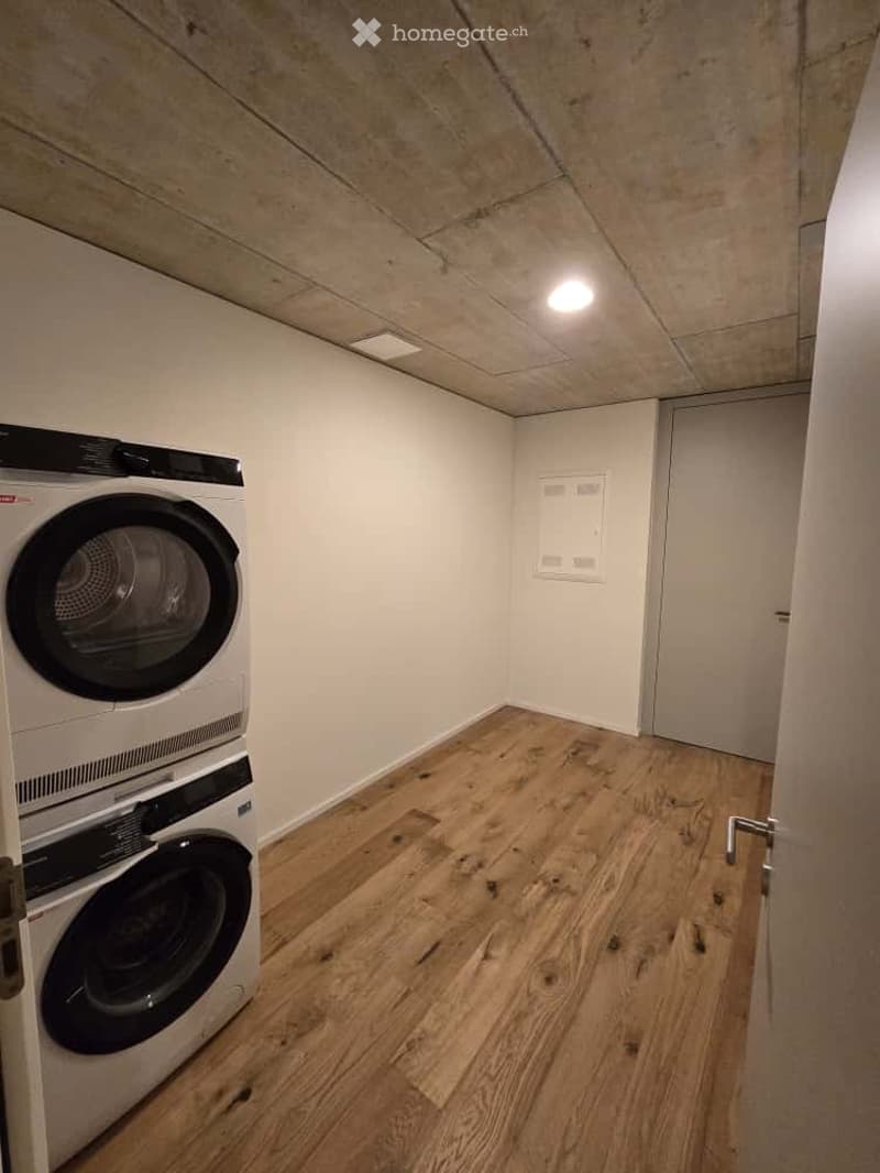 Furnished Room. A small, well-lit room with wooden flooring and white walls, featuring a stacked washing machine and dryer unit against one wall.
