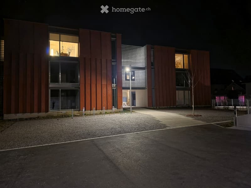 modern building, brown exterior, large windows, illuminated at night, gravel parking lot, street light