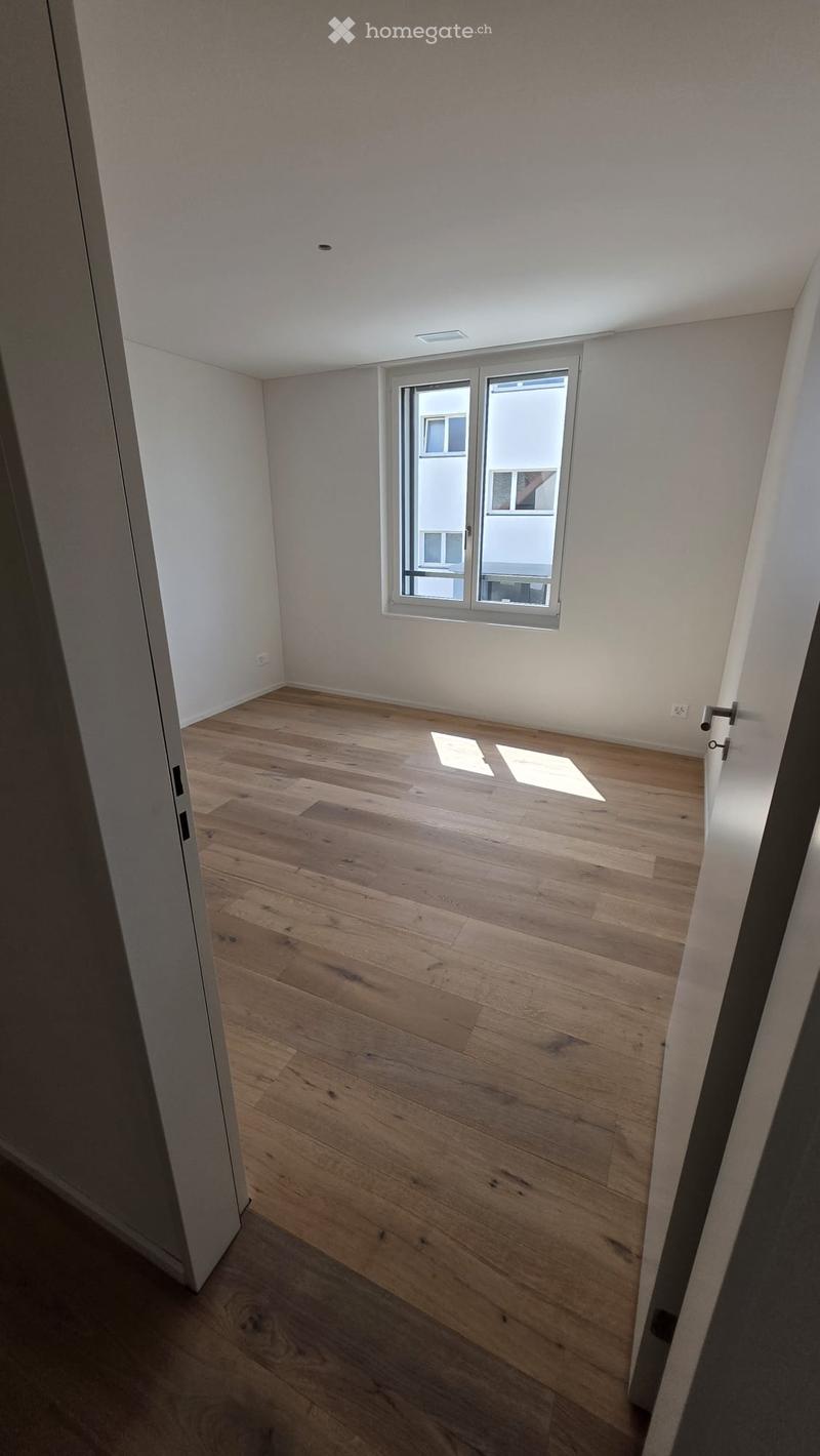 Empty room with light wooden flooring, white walls, and a large window letting in natural light.