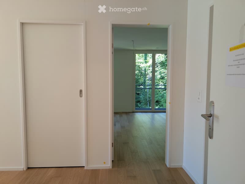 Empty hallway with wooden flooring, white walls, and a door leading to a room with large windows and a view of greenery.
