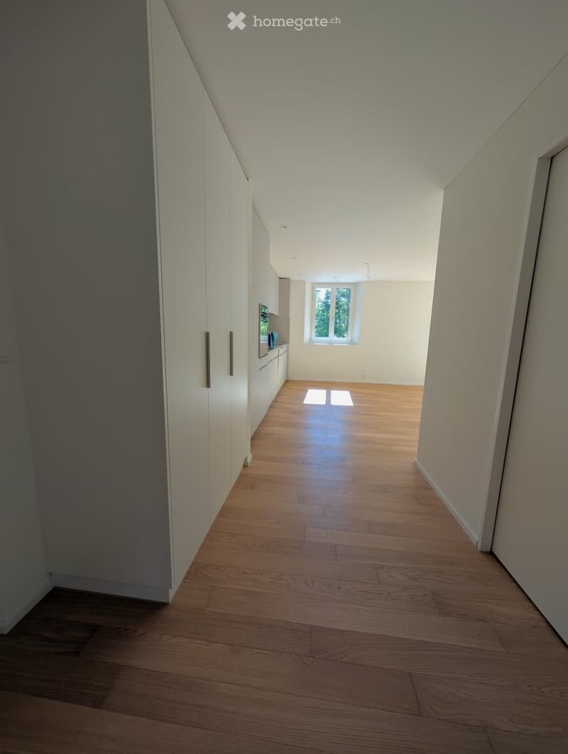 Empty hallway with wooden flooring, white walls, and a small window at the end, leading to a kitchen area.