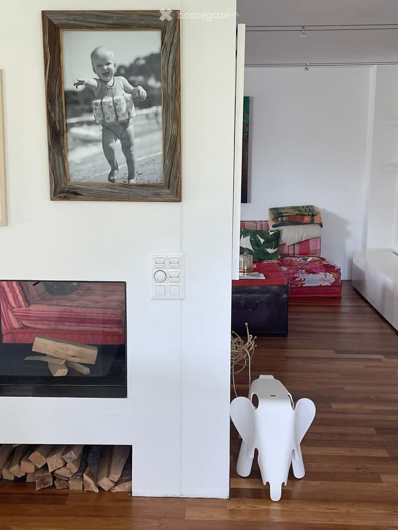 Fireplace with wooden logs, wooden floor, a white stool in the shape of an elephant, white walls, a picture of a child, two small couches