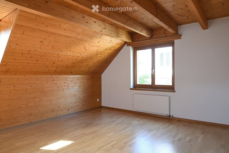 Empty attic room with wooden walls, ceiling beams, and hardwood floor. Features a large window with shutters and a white radiator below it.