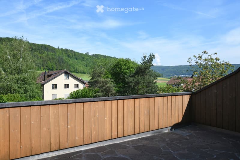 Wooden fence on a rooftop terrace overlooking a green, hilly landscape with houses and trees.