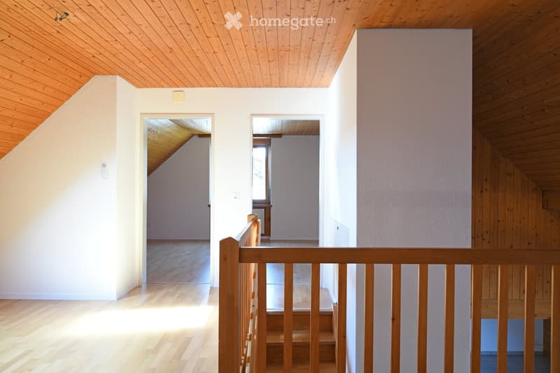 Bright, open hallway with wooden flooring and white walls, featuring a wooden railing and visible rooms beyond the doorways.