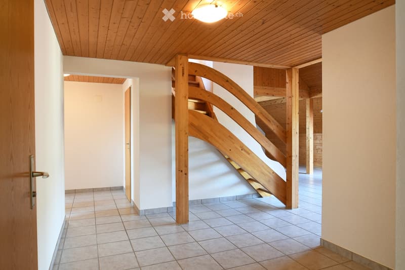 A modern hallway with wooden staircase, tiled floor, and light-colored walls. The ceiling features exposed wooden beams and a central light fixture.