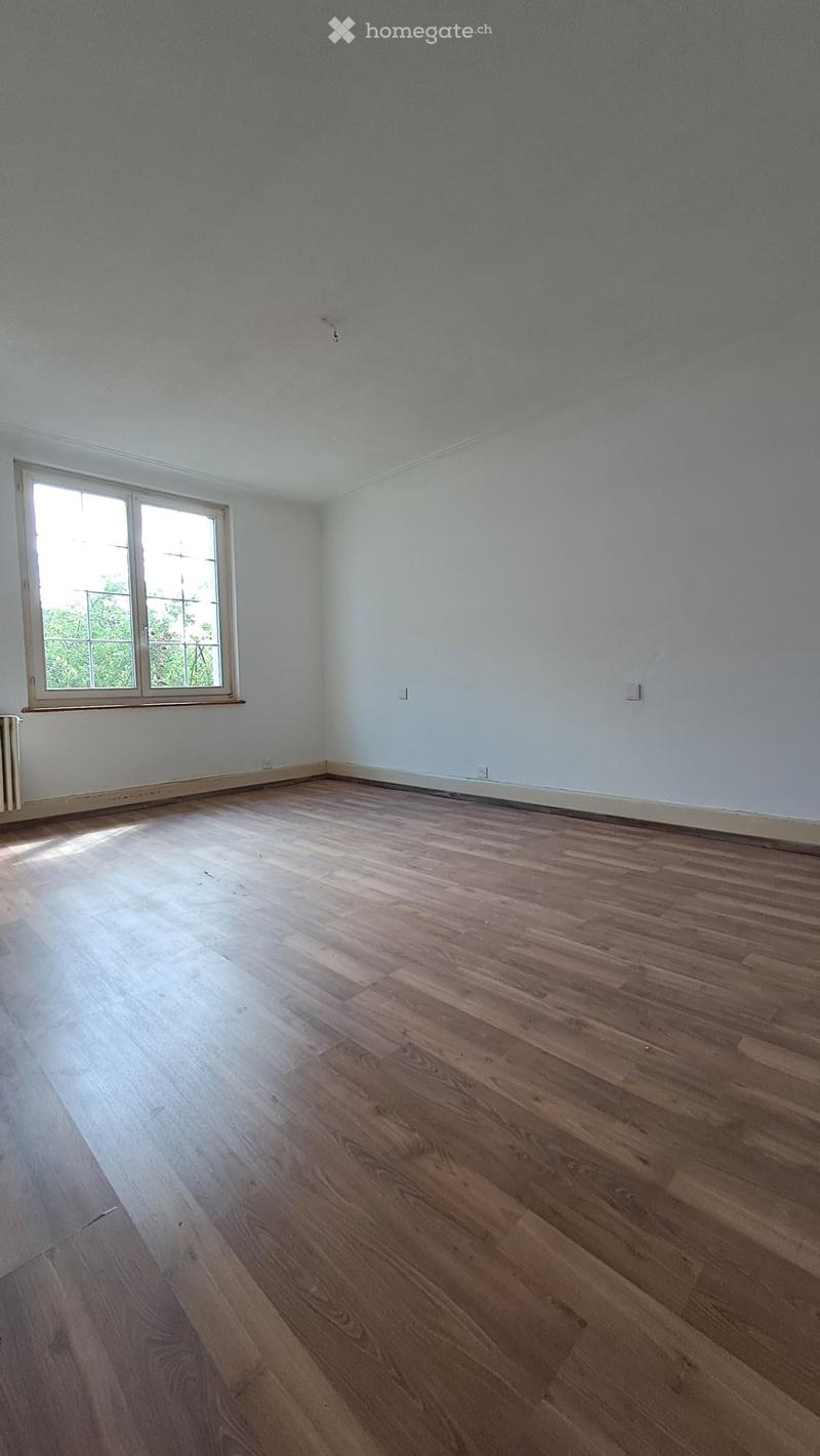 Empty room with wooden flooring and white walls, featuring a window with white frame and a radiator on the left side.
