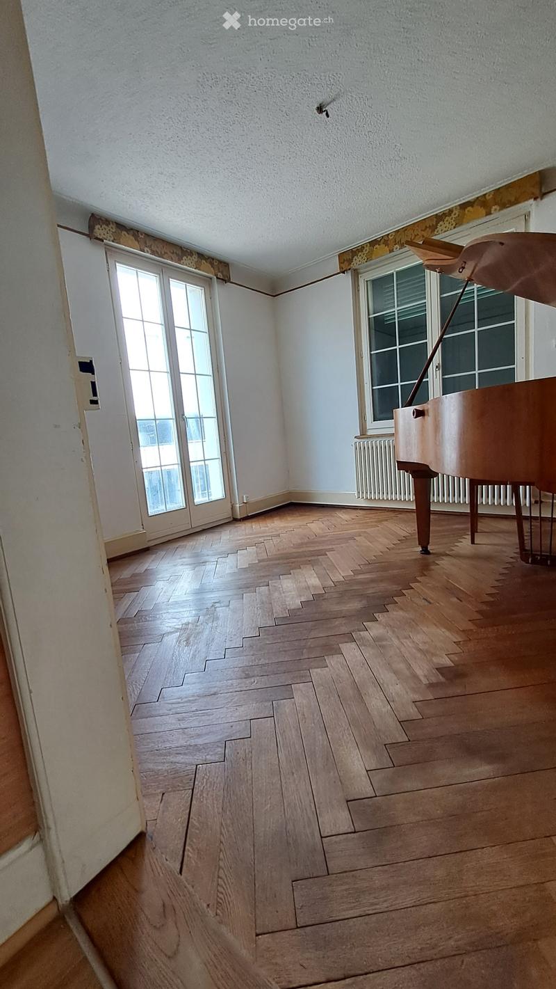 Empty room with wooden herringbone flooring, white walls, large windows, and a piano in the corner.