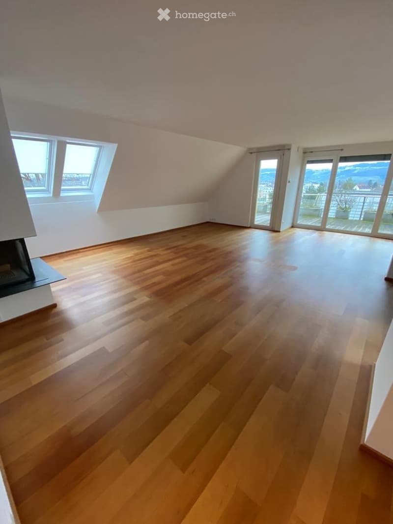 Bright, empty attic room with wooden flooring, large windows, and sliding glass doors leading to a balcony with a scenic view.