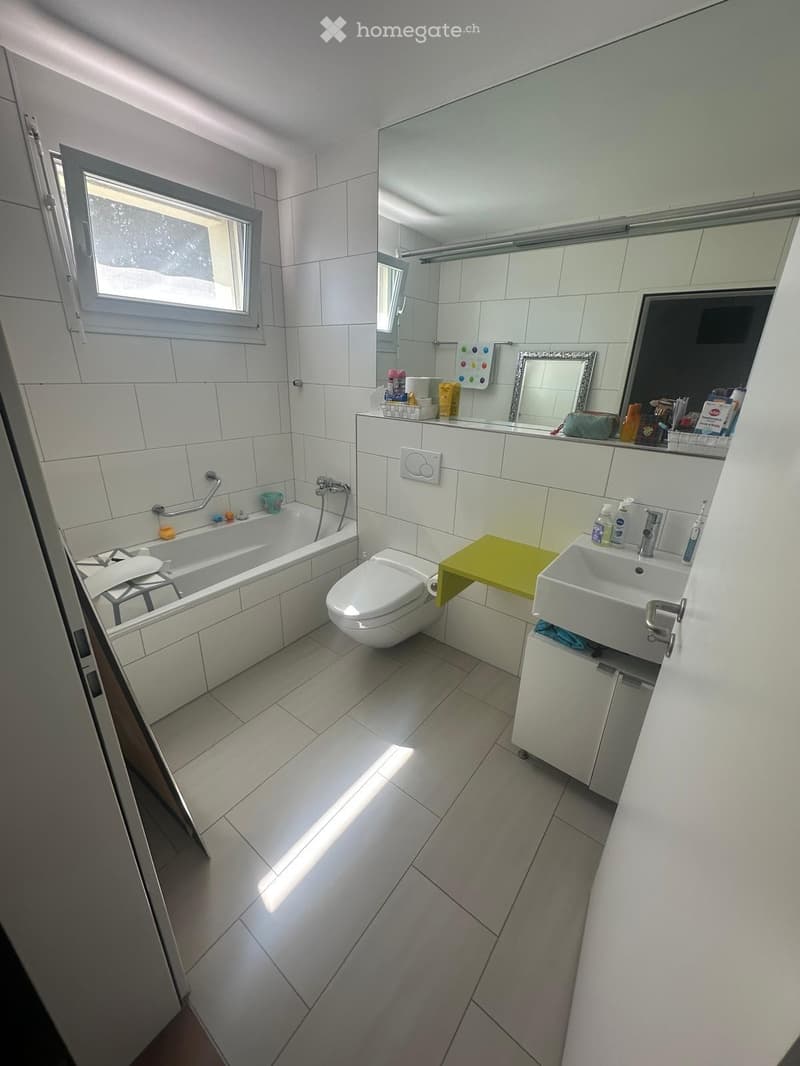 Modern bathroom with white tiles, bathtub, toilet, sink, mirror, and various toiletries on the counter.