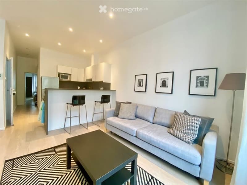 Modern open-concept living and dining area with a grey sofa, coffee table, and two stool chairs at a kitchen counter. The kitchen features white cabinets and a microwave. A floor lamp and framed art pieces are visible on the walls.