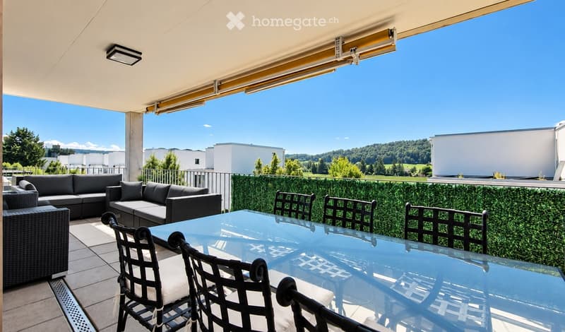 Balcony with a glass table, black chairs, and a grey sofa, overlooking a scenic landscape with trees and buildings.