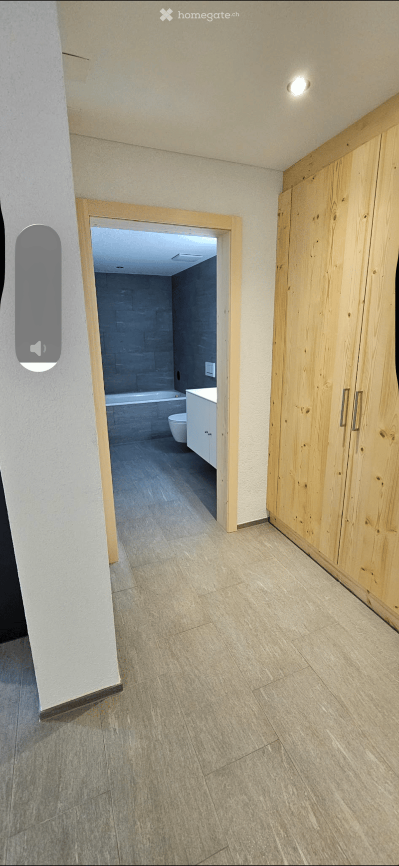 Empty hallway with tiled floor, light-colored walls, and a wooden wardrobe on the right side. An open doorway leads to a modern bathroom with a bathtub, toilet, and white cabinetry.