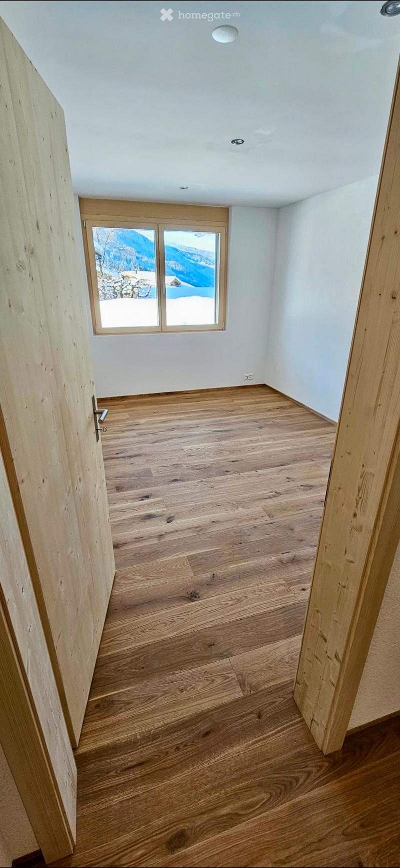 Empty room with wooden flooring, white walls, and a large window showing a snowy mountain view.
