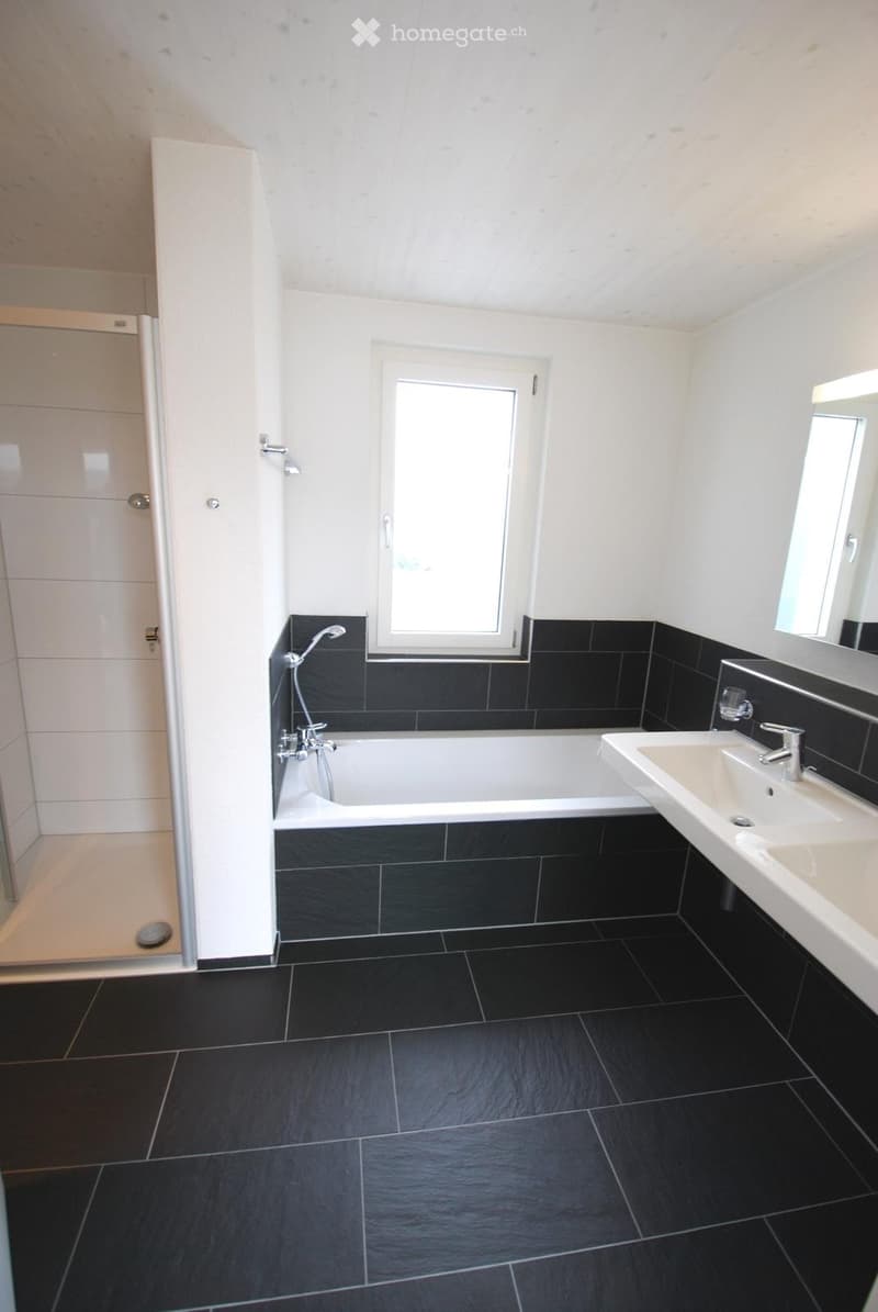 Modern bathroom with black tiled walls and floor, white bathtub, sink, mirror, and glass-enclosed shower area.