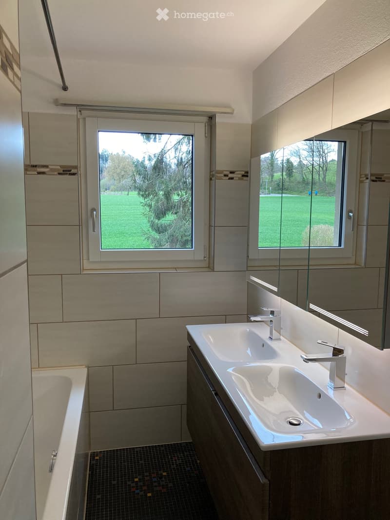 Modern bathroom with a double sink, bathtub, large windows offering a view of a green park, and sleek cabinetry.