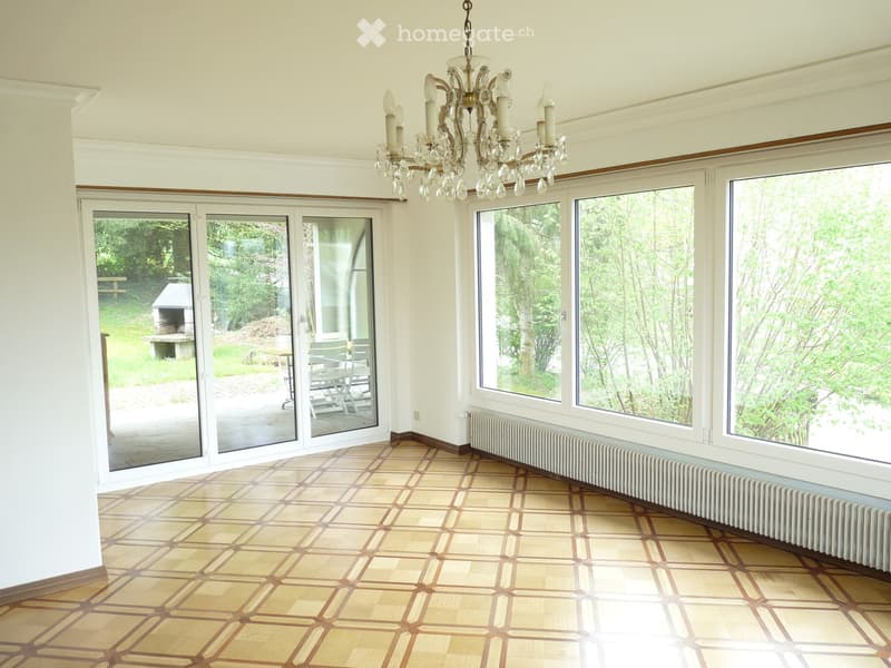 Bright, spacious room with large windows and sliding glass doors leading to an outdoor area. The floor is covered with a patterned parquet, and a chandelier hangs from the ceiling. The walls are painted in a light color, and a radiator is visible along on