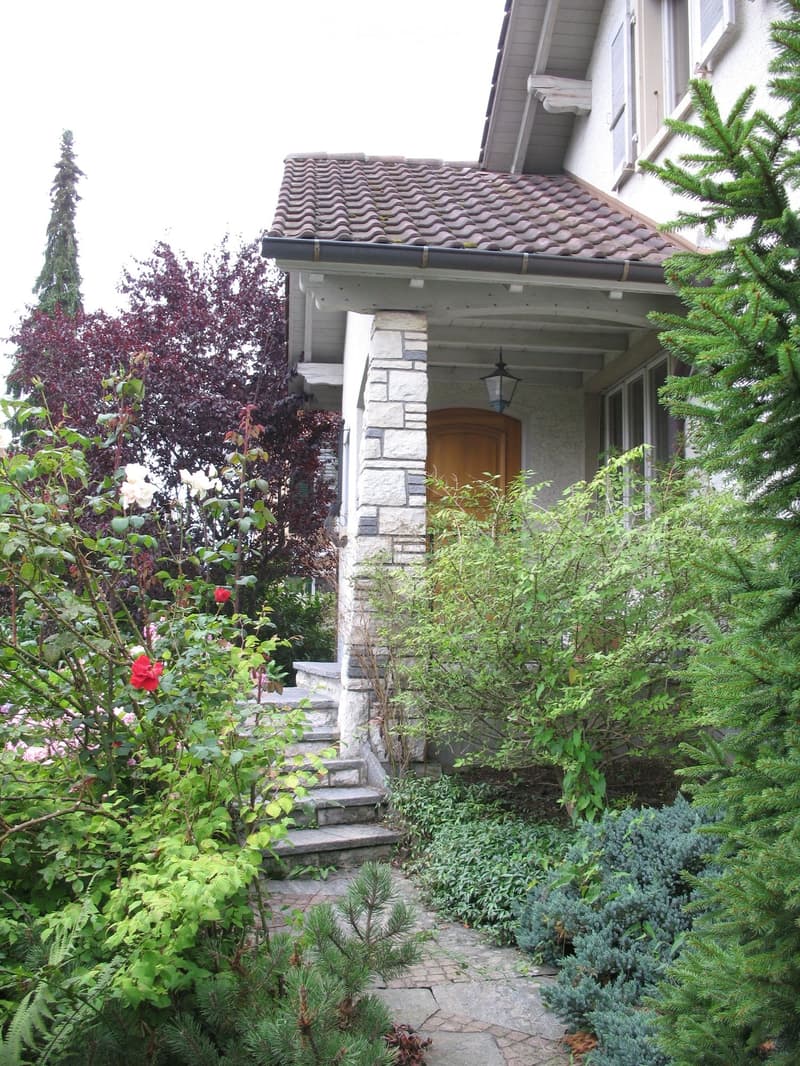 Two-story house with a tiled roof, stone entrance, and well-maintained garden with various plants and flowers.