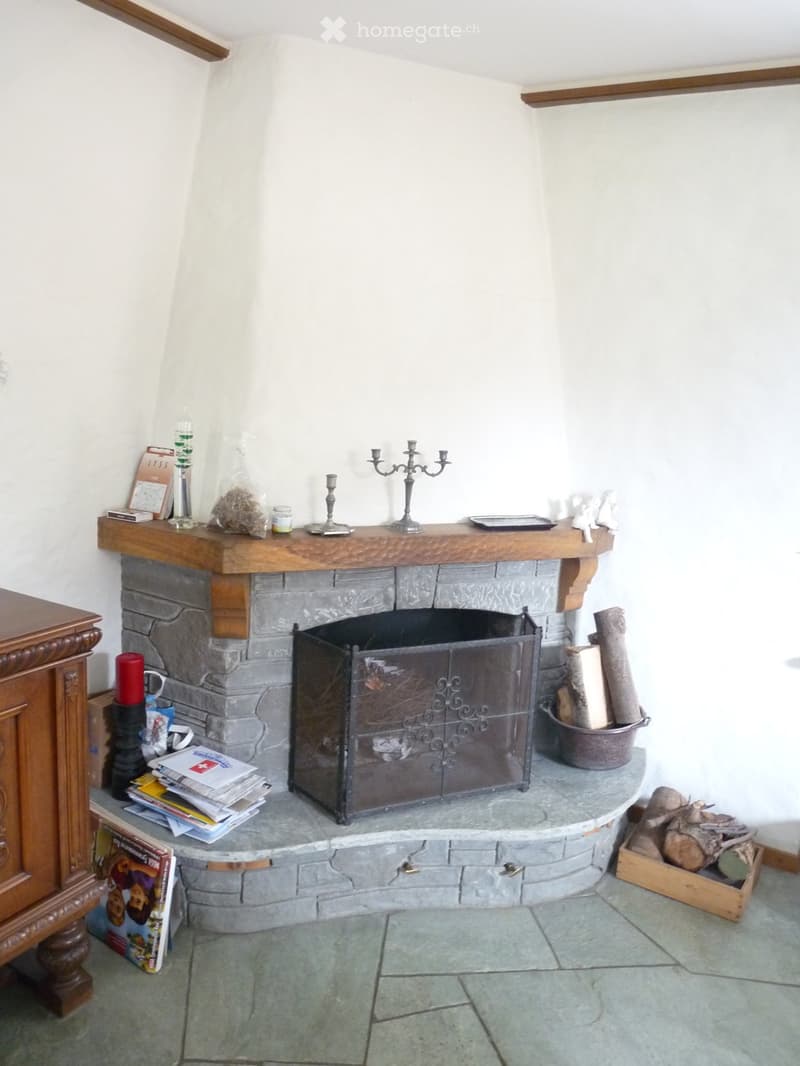 A stone fireplace with a wooden mantel, surrounded by decorative items including candles, a candle holder, a fire screen, and a stack of magazines. Wooden logs are stored nearby, and a wooden cabinet is visible to the left.