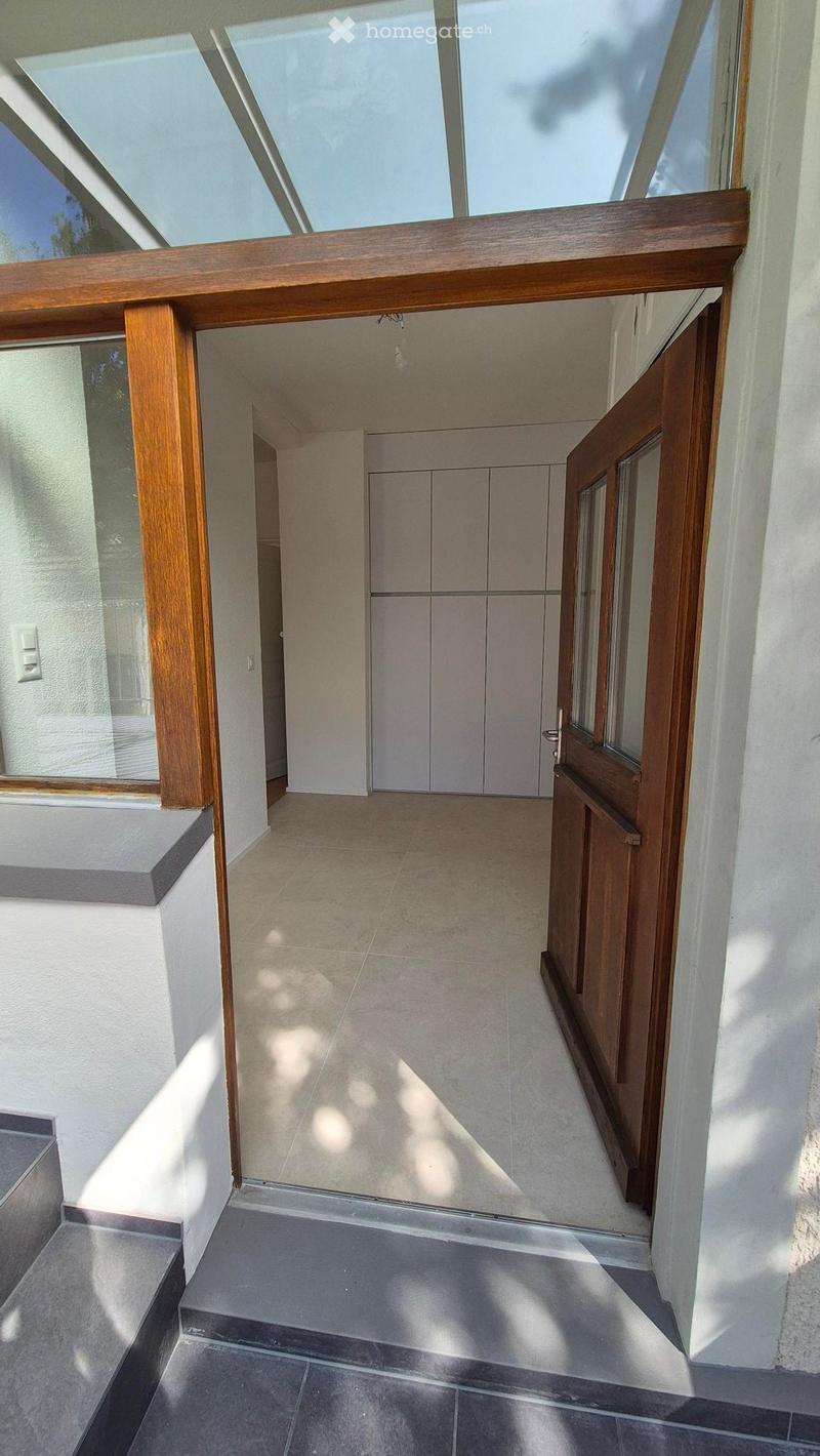 Bright, modern hallway with tiled floors, white walls, and a wooden door leading to a room with built-in cabinets.