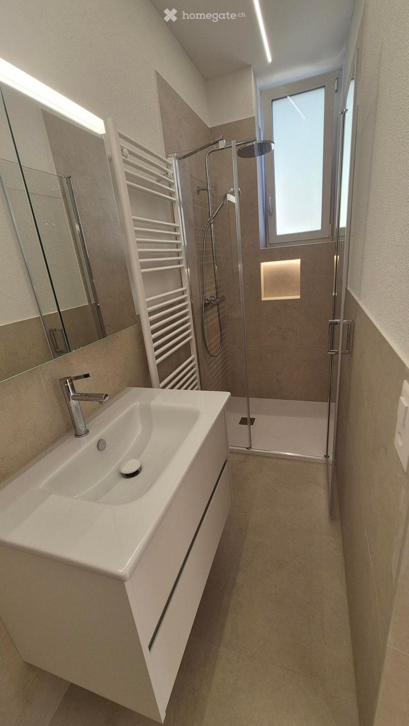 Modern bathroom with a white sink, chrome faucet, large mirror, glass-enclosed shower with a modern showerhead, and a towel rack. The space features beige tiles and a window for natural light.