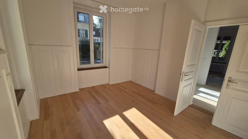 Empty room with wooden flooring, white walls, and a large window with a built-in bench. An open door leads to another room.