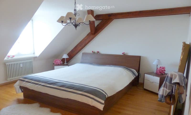 Cozy attic bedroom with wooden beams, slanted ceilings, and large windows. Features a wooden bed with striped bedding, two nightstands with lamps, and a radiator under the window.