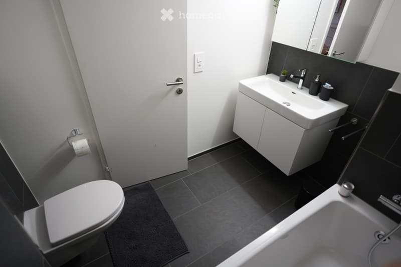 Modern bathroom with white toilet, sink, and bathtub, black tiles, and a large mirror above the sink.