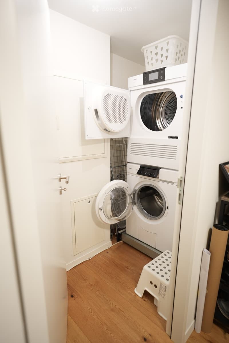 Compact bathroom with a washing machine and dryer unit, wooden flooring, and a small white stool.