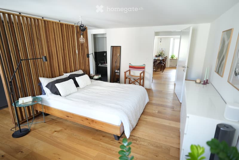 Modern bedroom with wooden flooring, a large bed with white and black bedding, a nightstand, a chair, and a mirror. The room features a wooden wall panel and a view into an adjacent dining area.
