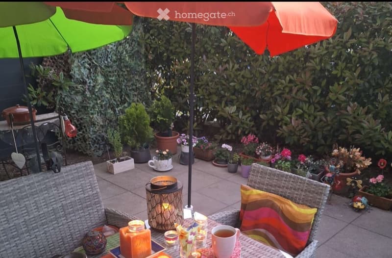 A cozy outdoor patio area with wicker chairs, colorful cushions, a table with candles and a teacup, surrounded by potted plants and flowers, with colorful umbrellas providing shade.