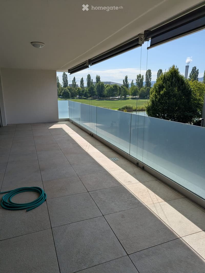Balcony with tiled floor, glass railing, and a view of a park and water body.