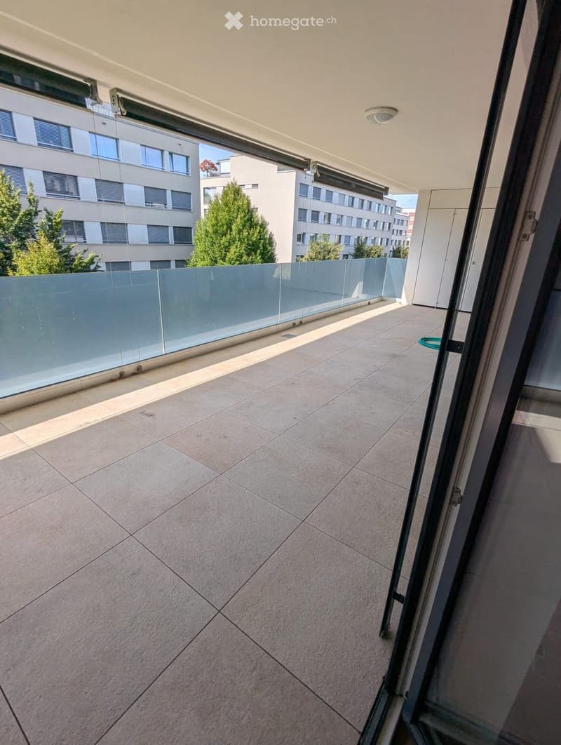 Balcony with tiled floor, glass railing, and view of surrounding buildings and greenery.