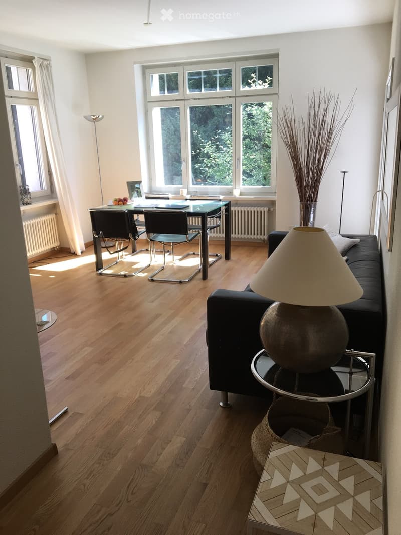 Bright living room with wooden flooring, large windows, sofa, coffee table, dining table, chairs, and decorative vase.