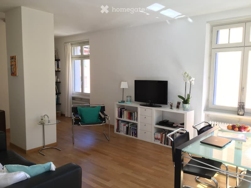 Bright living room with wooden flooring, large windows, a black sofa with cushions, a glass coffee table, a bookshelf with books and a TV, and a glass dining table with chairs.