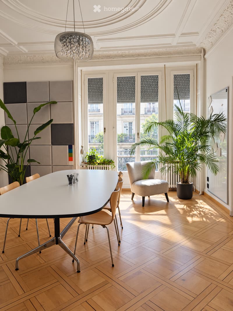 Elegant dining area with a large round table, modern chairs, potted plants, and a chandelier, featuring wooden herringbone flooring and large windows with views.
