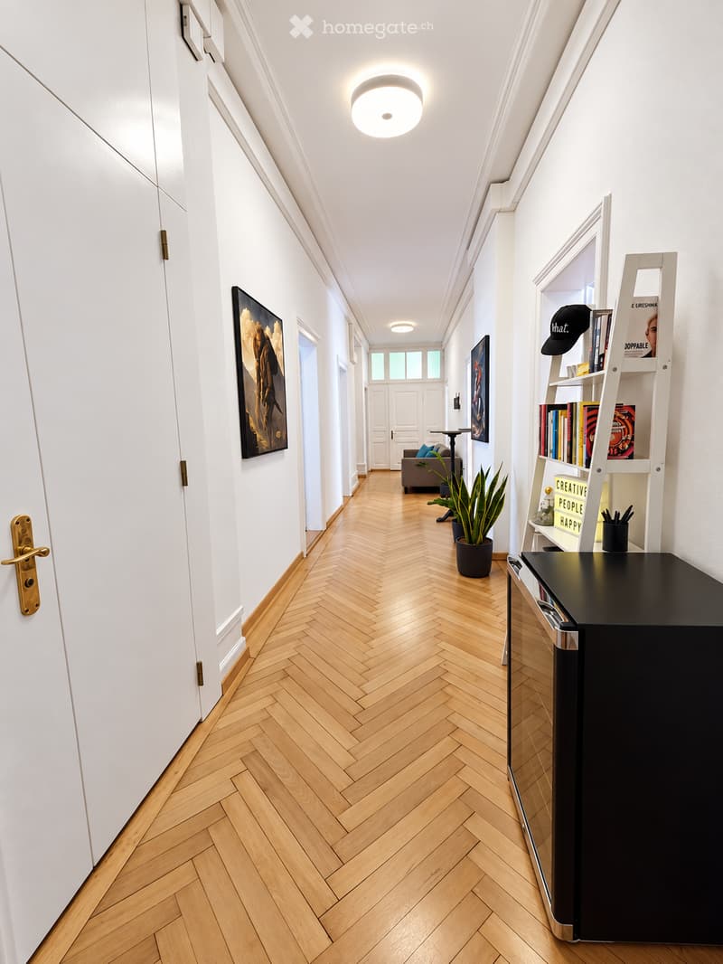 A well-lit hallway with herringbone wooden flooring, white walls, and modern decor including a bookshelf, potted plant, and artwork.