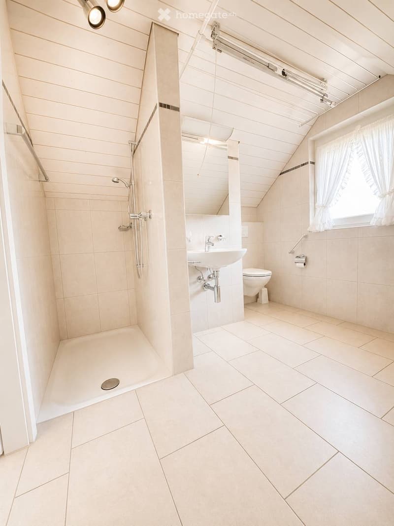 Modern bathroom with a walk-in shower, a sink, and a toilet. The space features light-colored tiles and a sloped ceiling with built-in lighting.