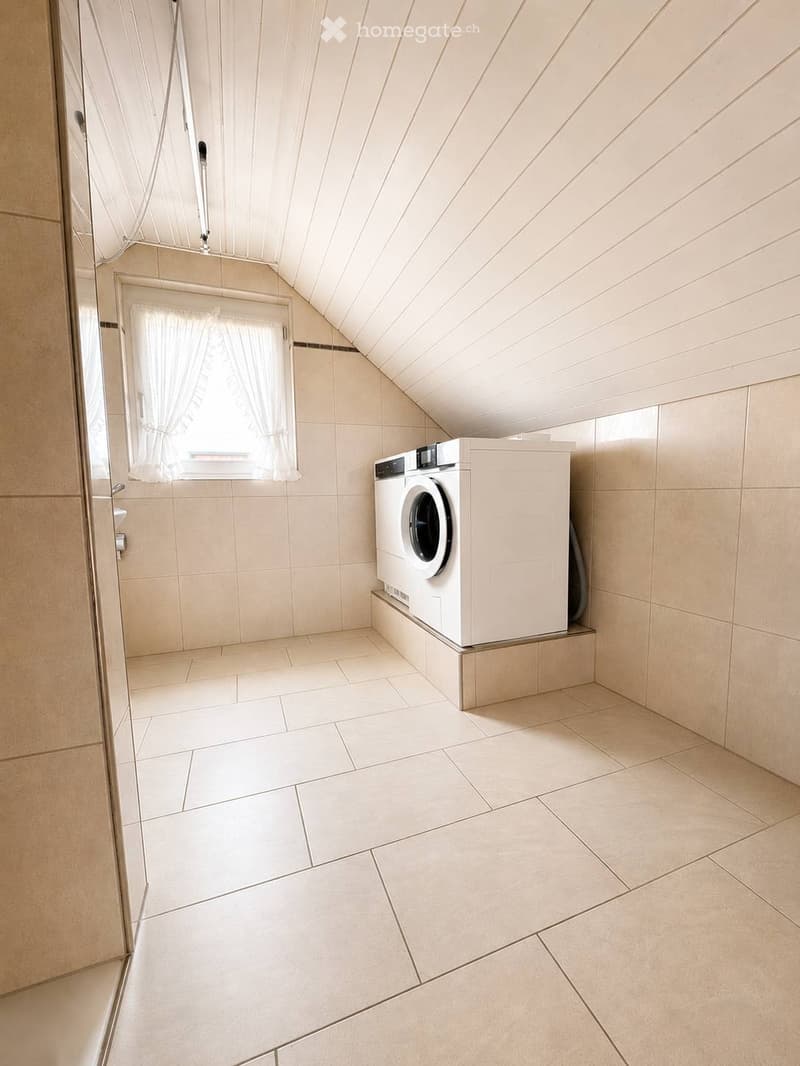 Clean, modern bathroom with a washing machine, tiled walls and floor, and a window with white curtains.