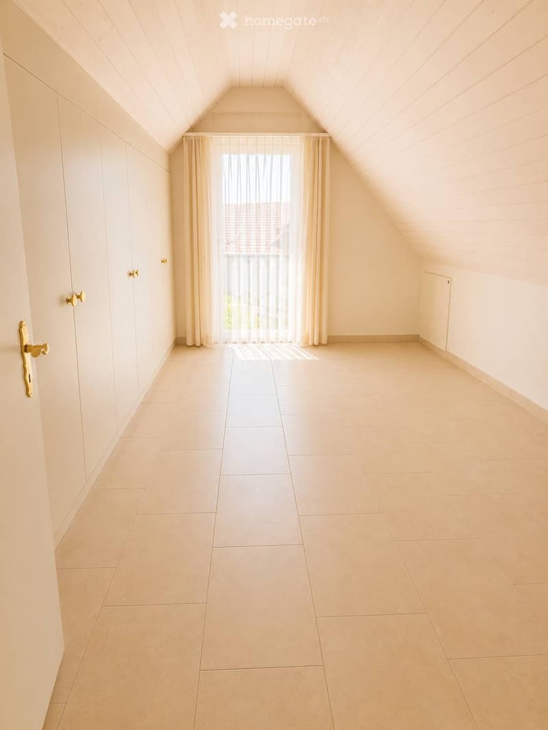 Empty attic room with slanted ceilings, tiled floor, large window with sheer curtains, and built-in wardrobes.