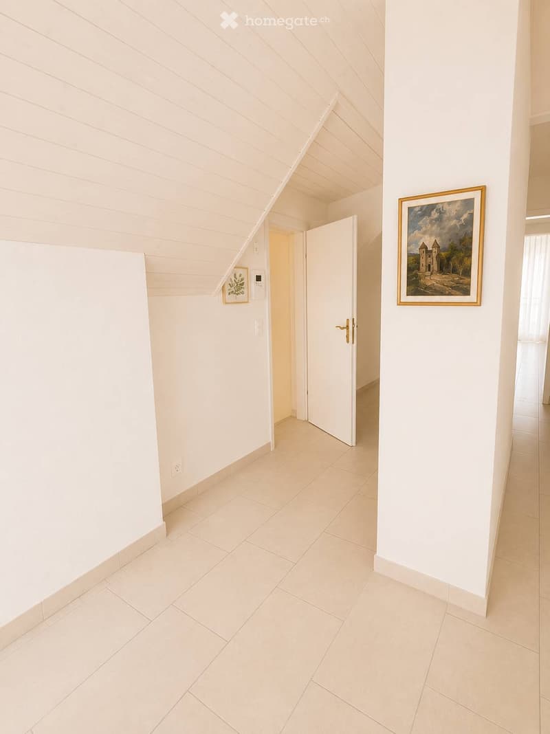 Clean, minimalist hallway with white walls, tiled floor, and two framed pictures. An open door leads to another room.