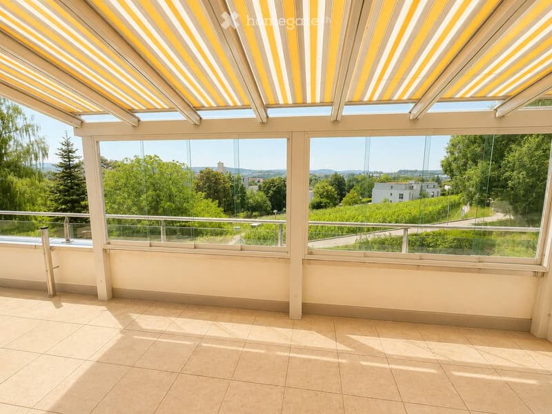 Bright, modern balcony with large glass windows and a retractable striped awning, offering scenic views of greenery and distant buildings.