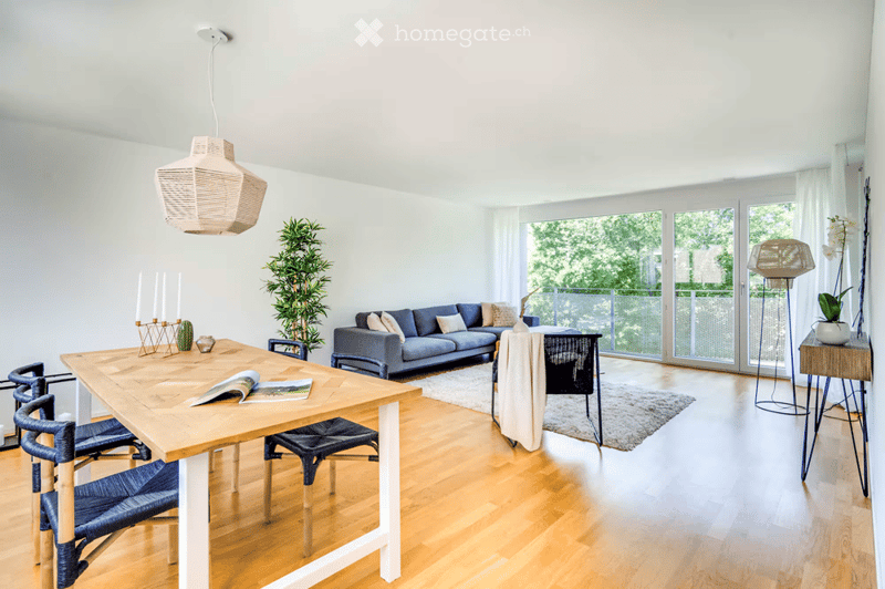 Bright living room with wooden flooring, large windows, and a balcony. Features a blue sectional sofa, wooden dining table with chairs, floor lamp, and decorative plants.