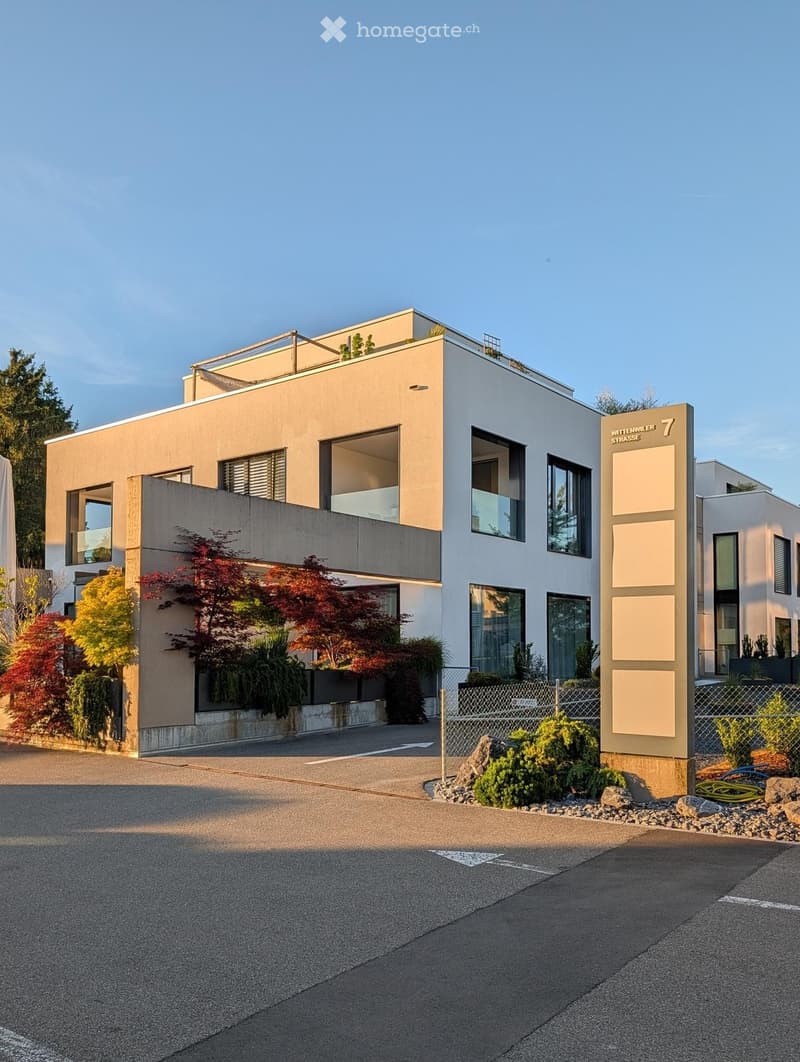 Modern two-story building with a flat roof, large windows, and a balcony on the upper level. The building has a combination of beige and white exterior walls and is surrounded by landscaped greenery.