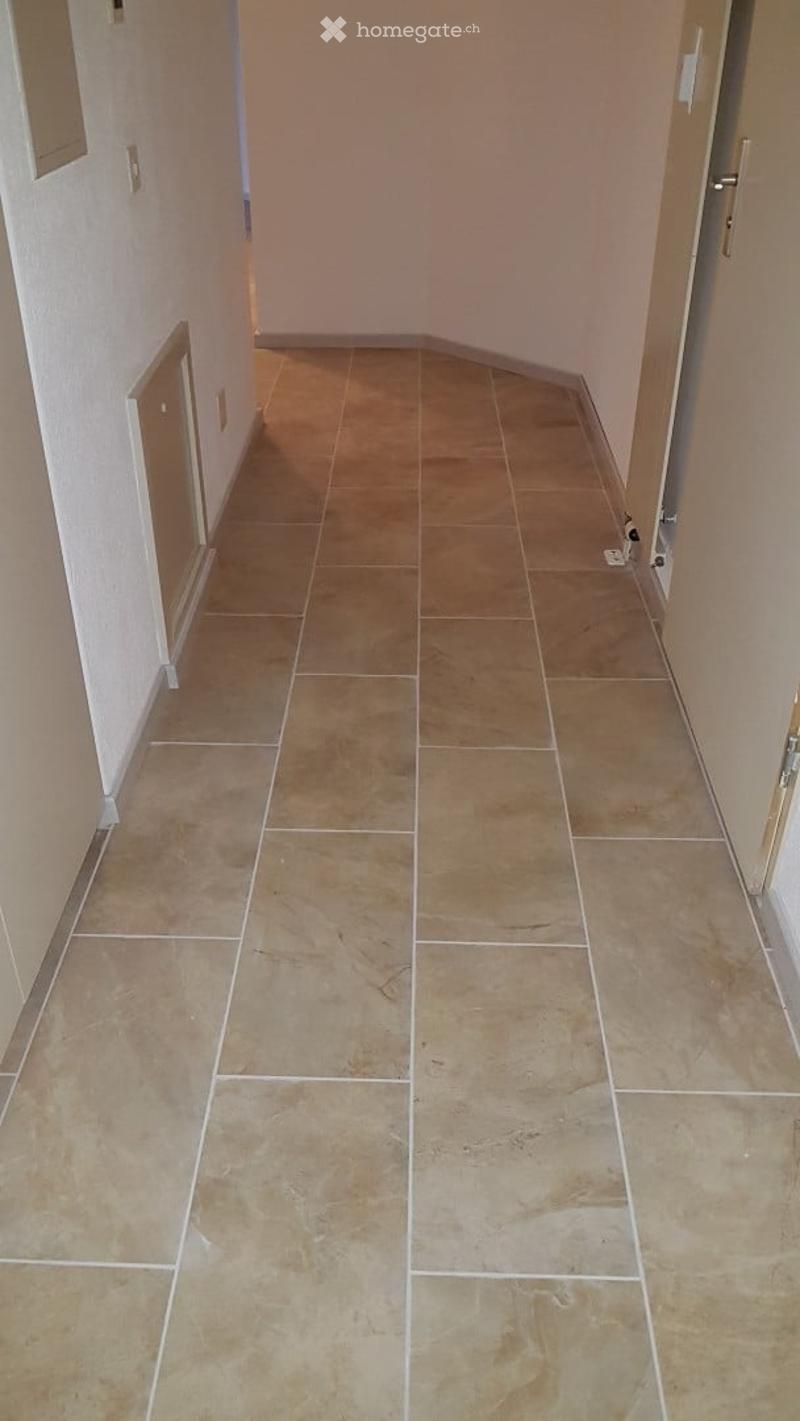 A clean, tiled hallway with beige tiles and white grout lines, featuring light-colored walls and a door at the end.