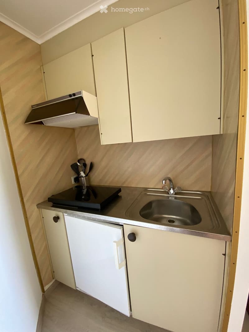 Compact kitchen with light-colored cabinets, a stainless steel sink, a stove, and a range hood. The countertop is dark, and utensils are placed near the stove.