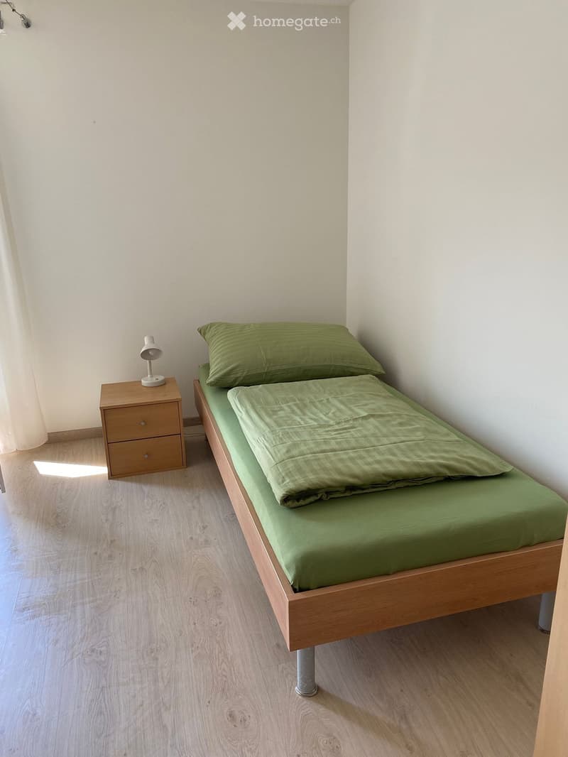 A small, neatly arranged bedroom with a single bed, green bedding, a wooden nightstand, and a lamp.